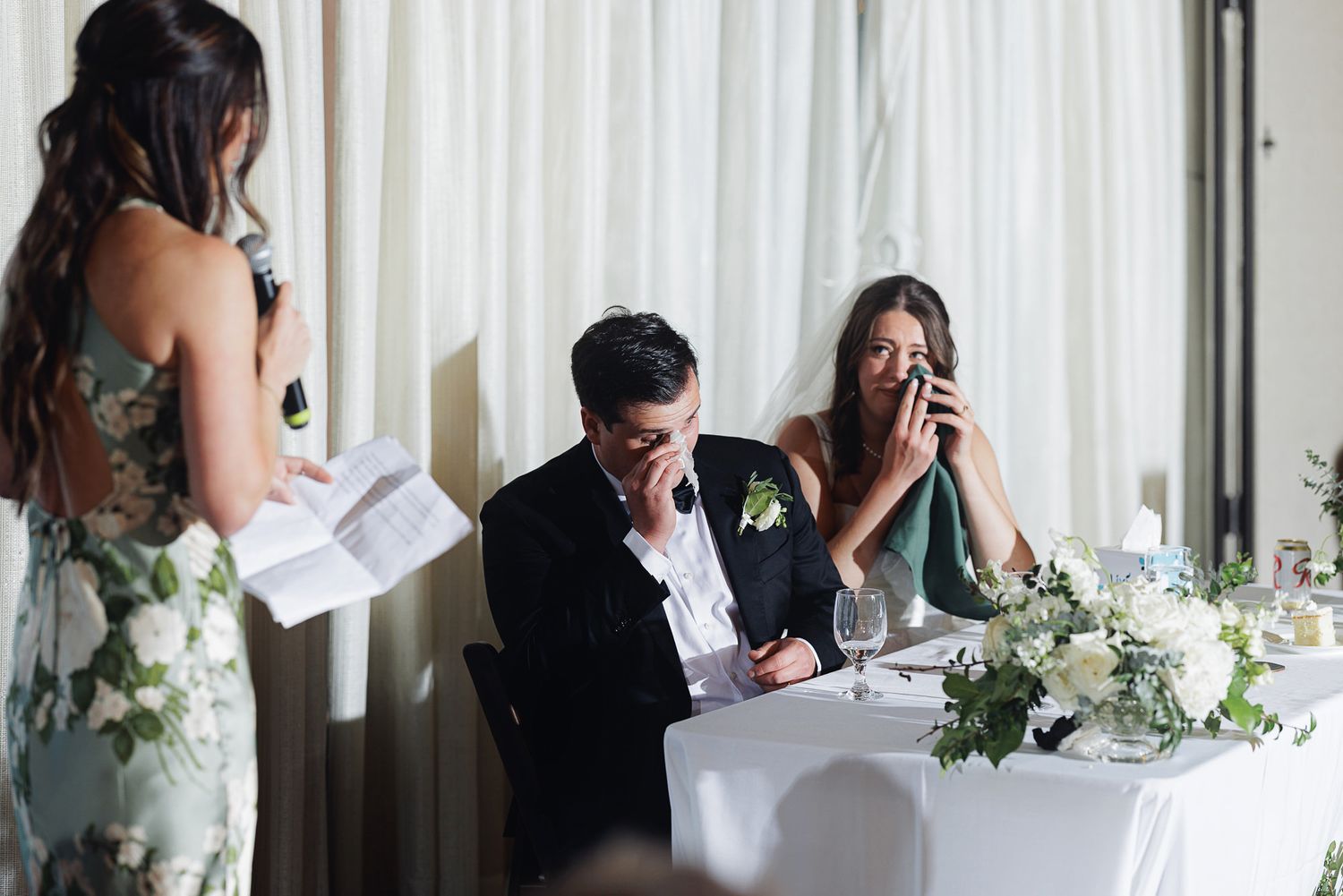 Emotional bride and groom crying during heartfelt wedding speech at elegant white floral reception table.