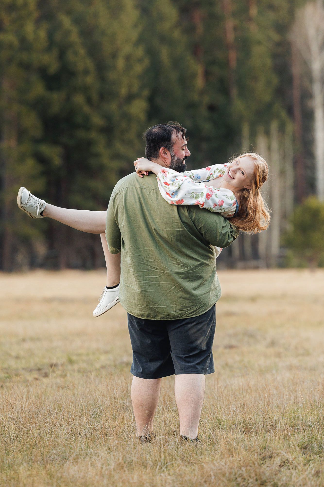 Man carrying laughing woman in floral dress across grassy field with pine trees in background.