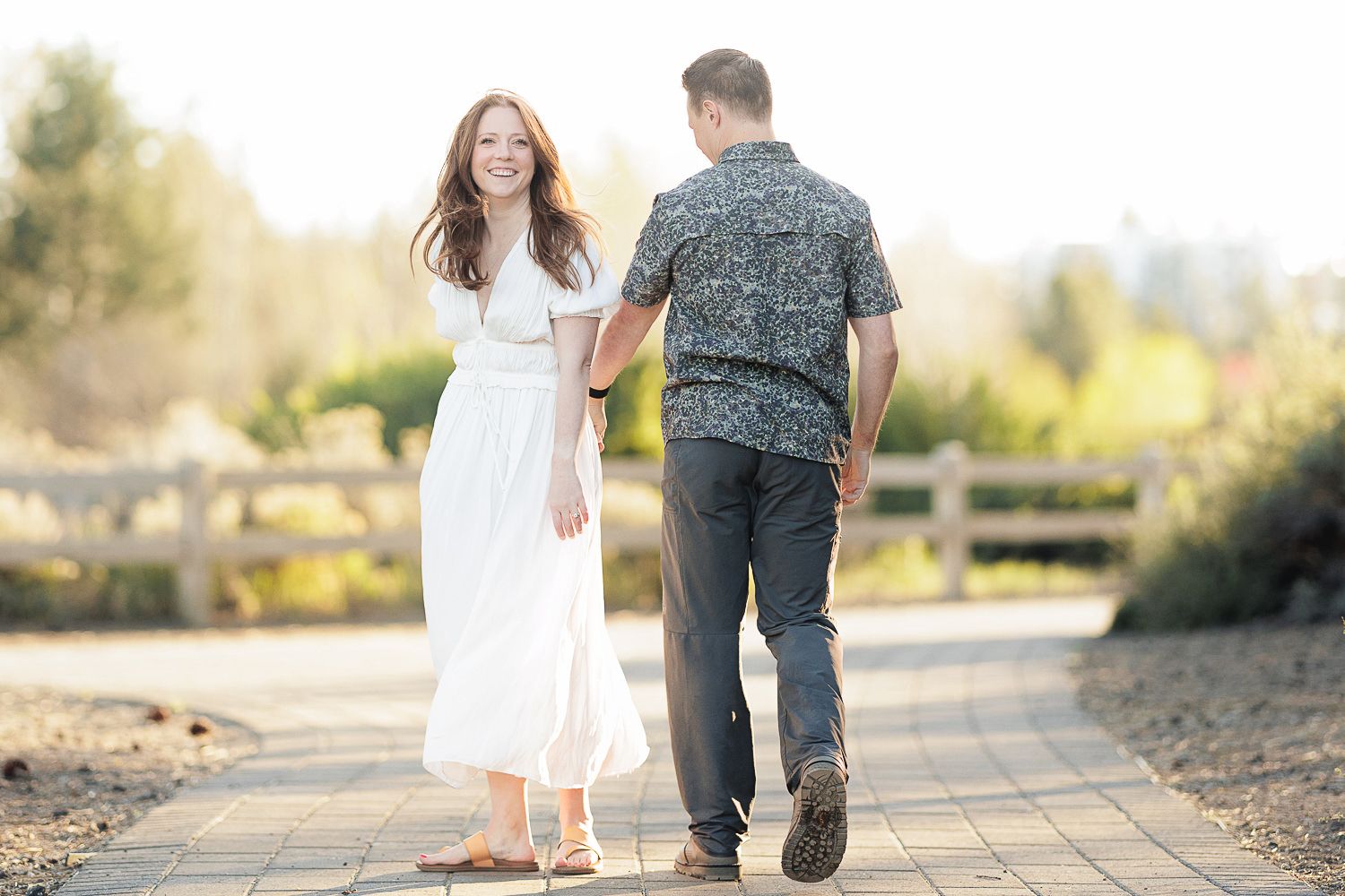 A couple holds hands while walking along a sunlit pathway in a beautiful outdoor setting.