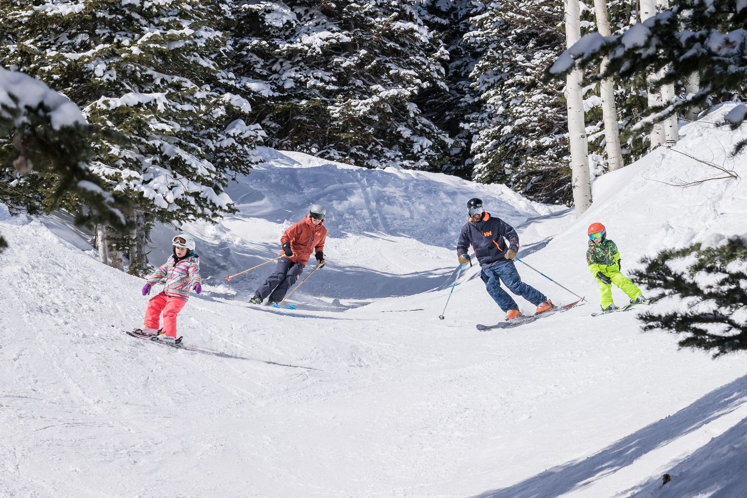 Four skiers carving down a snowy mountain slope between evergreen trees on a bright winter day.