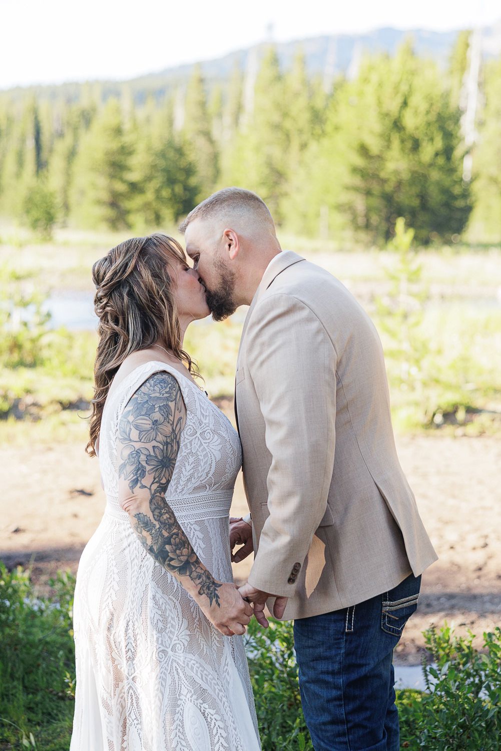 A couple in wedding attire share a tender moment outdoors with evergreen trees and mountains in the background.