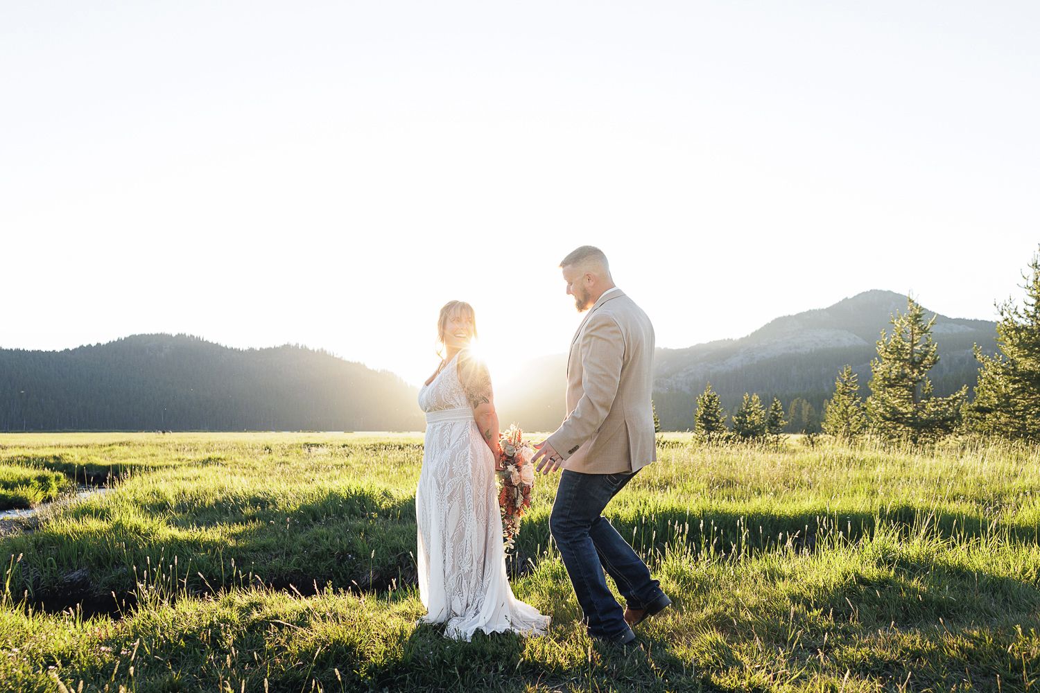 Newly married couple walks hand in hand through a sunlit mountain meadow during golden hour.