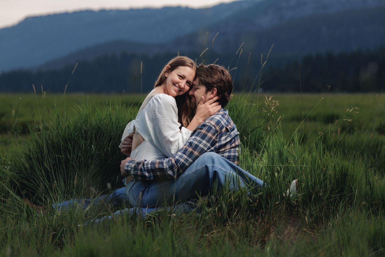 A romantic couple sits together in a green meadow during sunset in the mountains.