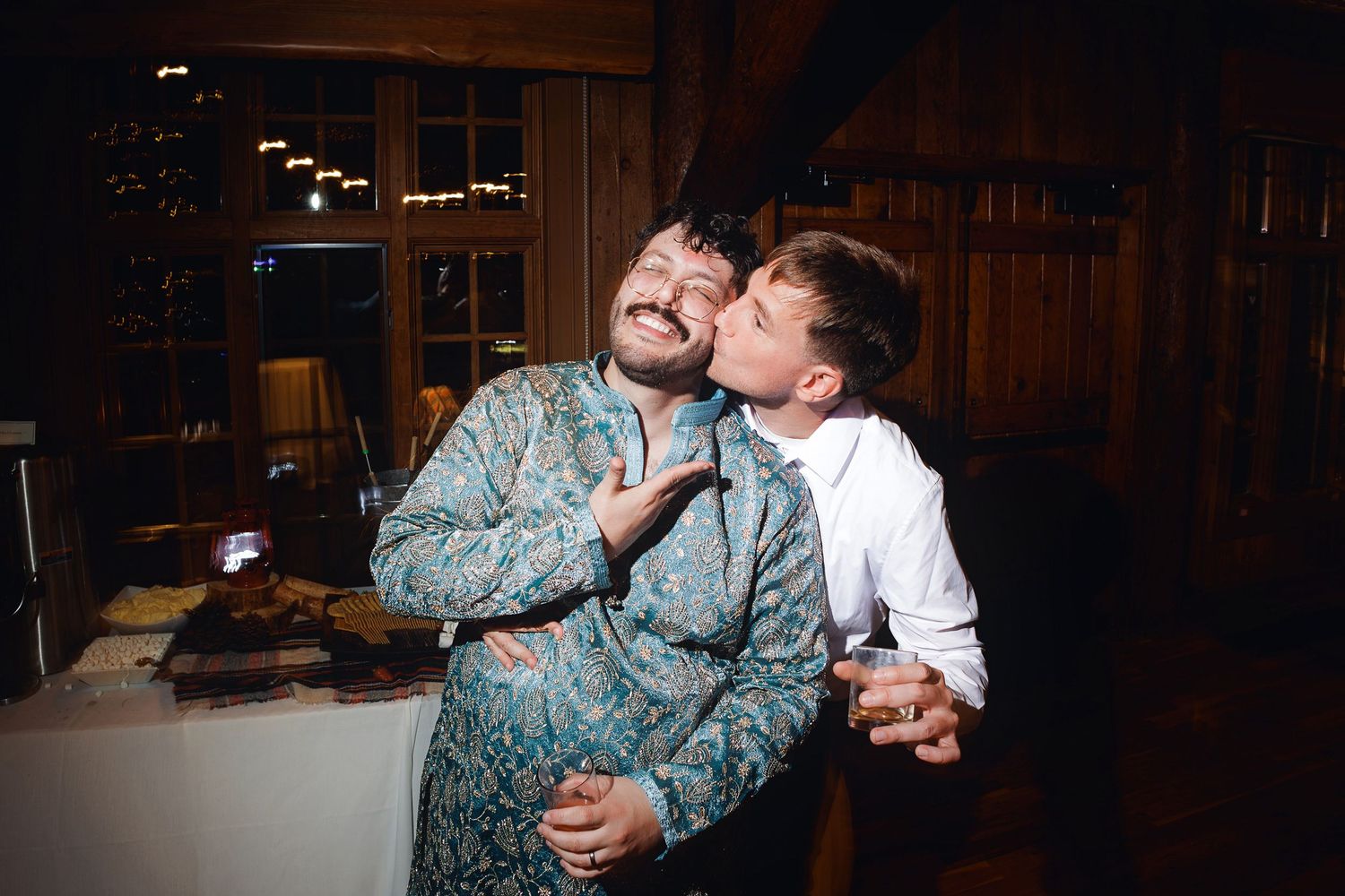 Two men celebrating at a party, one kissing the other on the cheek while making a peace sign indoors.