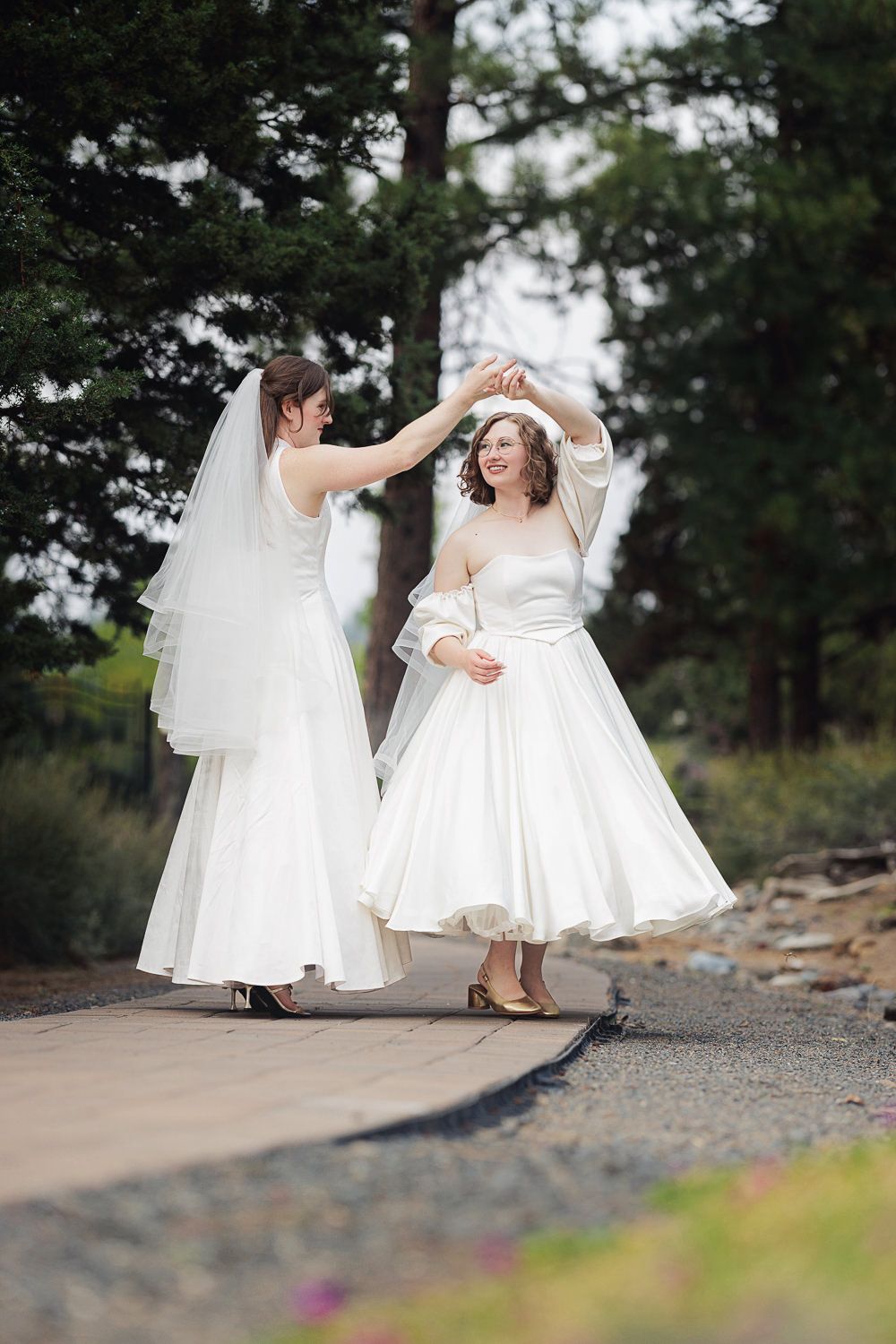 Two people in white wedding dresses dance and twirl together on a rocky outdoor path surrounded by greenery.