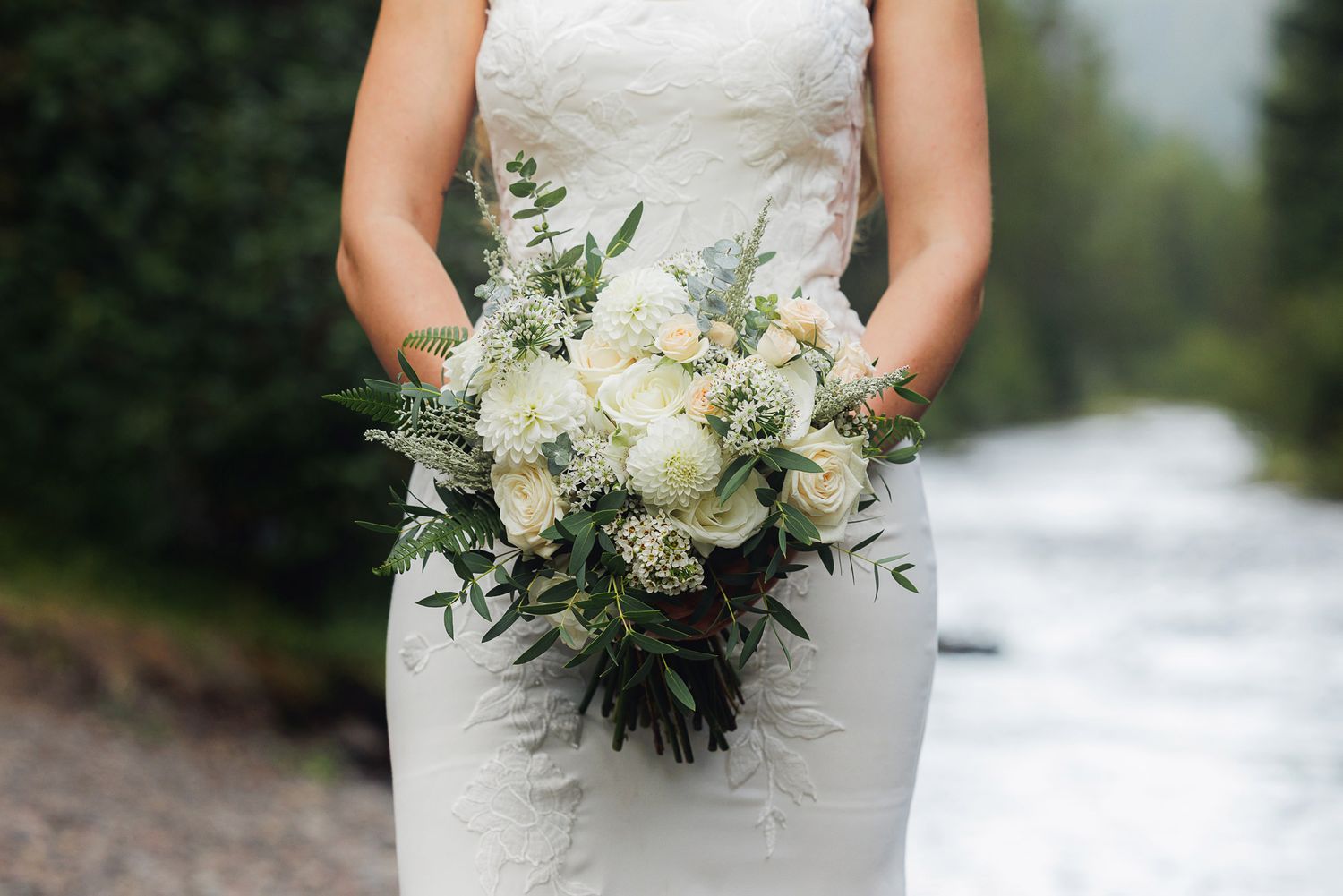 A bride holds an elegant white and green floral bouquet against her lace wedding dress in an outdoor setting.