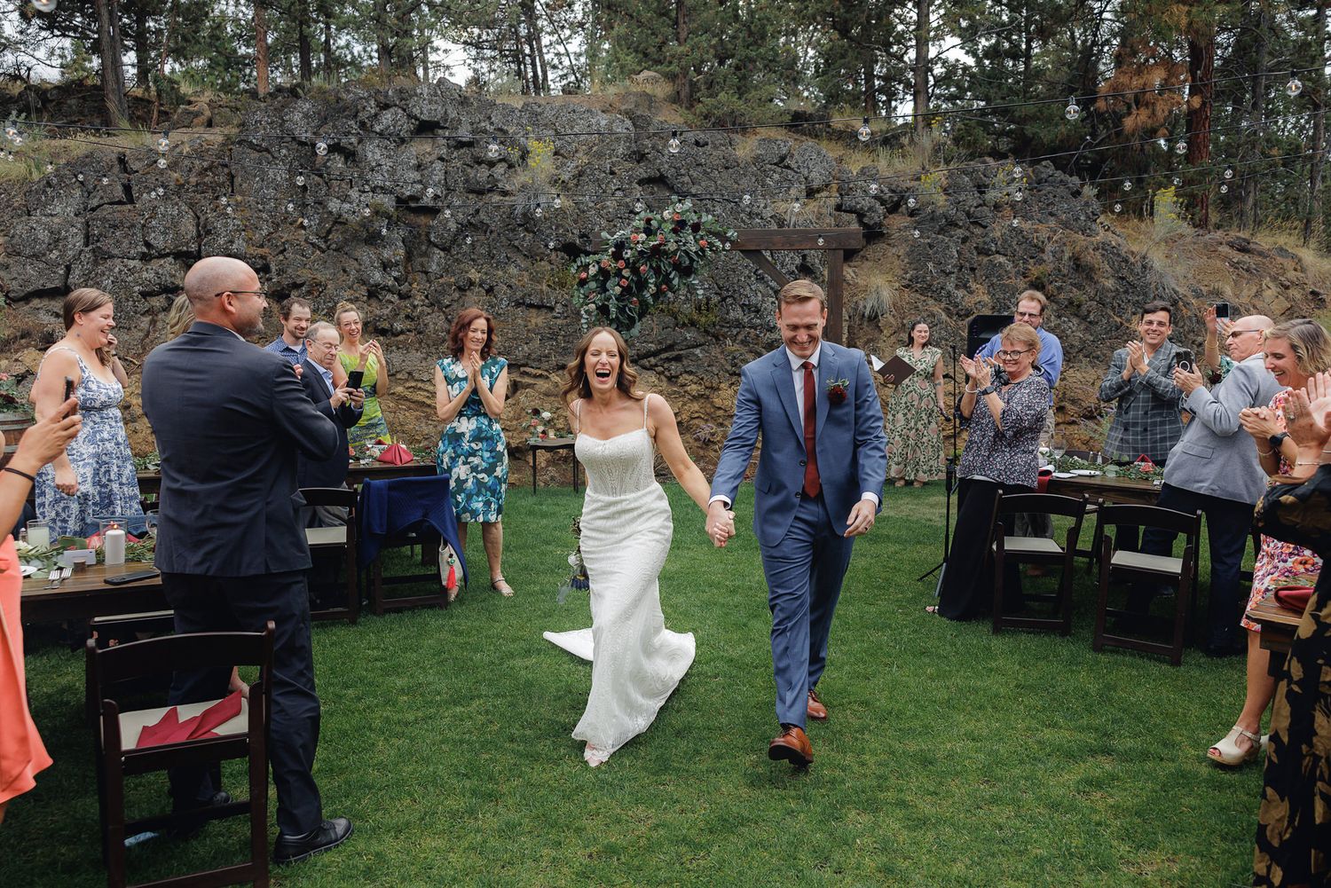 Wedding celebration on a grassy lawn with rocky backdrop as guests watch a couple make their entrance.