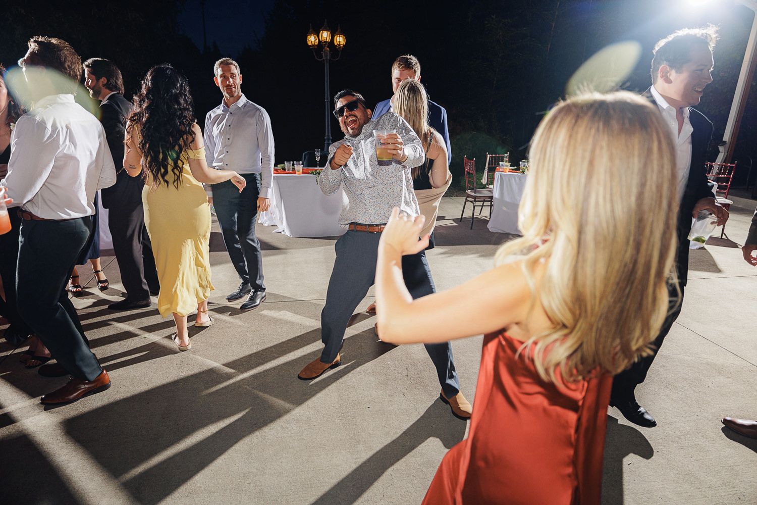 Wedding guests dance and celebrate at an evening outdoor reception with dramatic lighting and shadows on the dance floor.