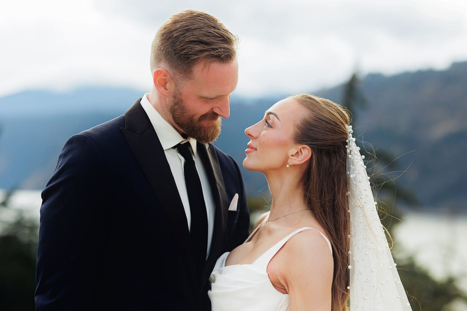 A bride and groom share a romantic moment outdoors with mountain views, she wearing a veil and he in a navy suit.