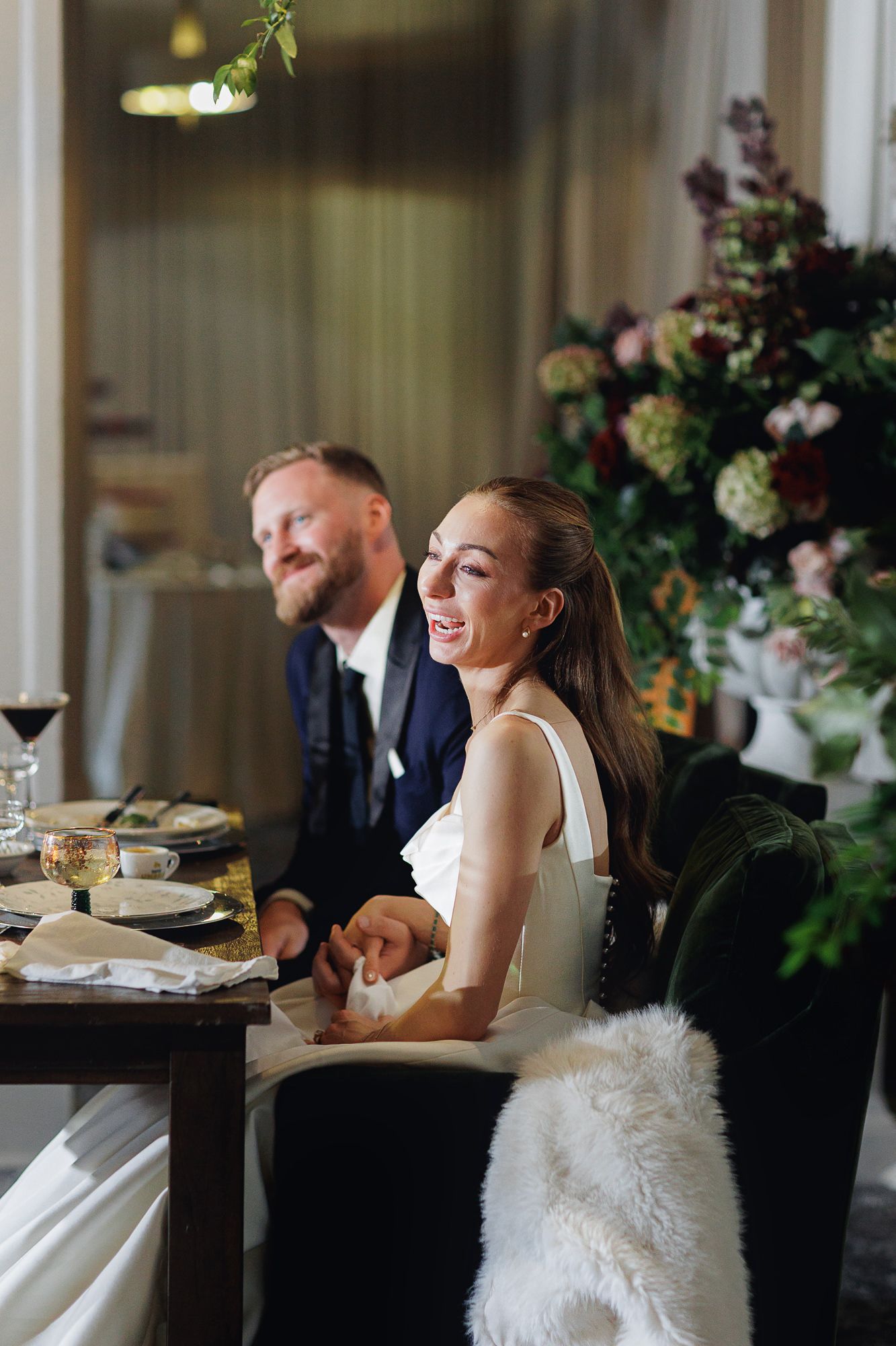 Bride and groom laughing at wedding reception table with floral arrangements and elegant decor.