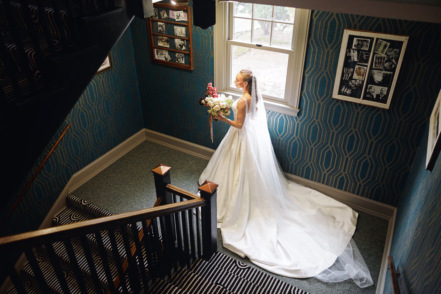 Bride in white gown and veil holding bouquet stands at top of staircase in elegant blue wallpapered room.