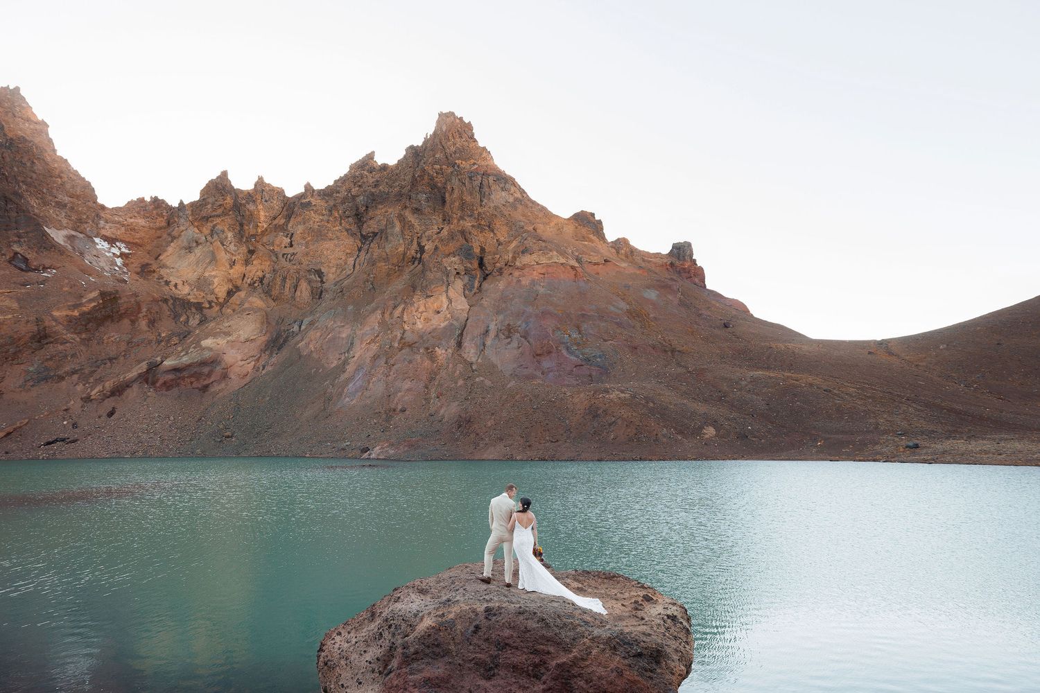 Bride and groom in wedding attire stand on a large rock beside a turquoise mountain lake beneath rugged reddish-brown peaks.