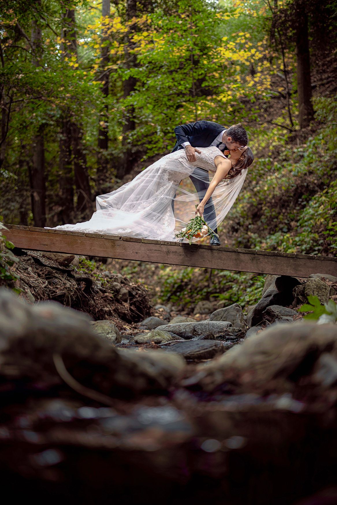 A romantic wedding photo of a couple sharing a dramatic dip pose on a wooden footbridge in a lush forest setting.
