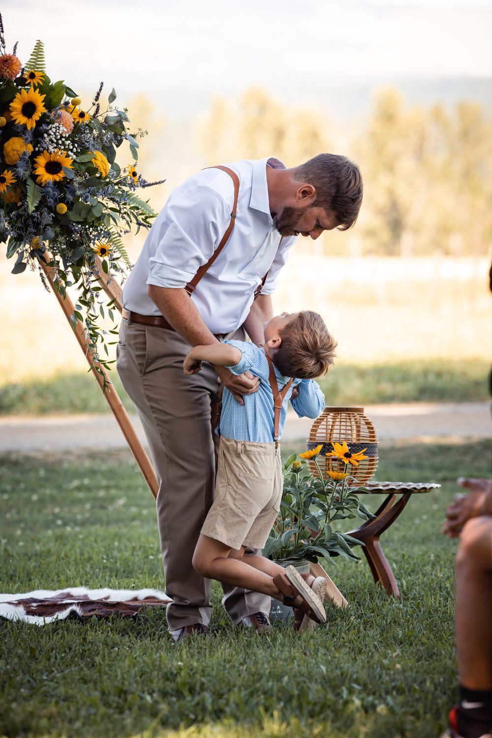 A tender outdoor family moment next to a rustic wooden stand with sunflowers and a picnic basket in a countryside setting.