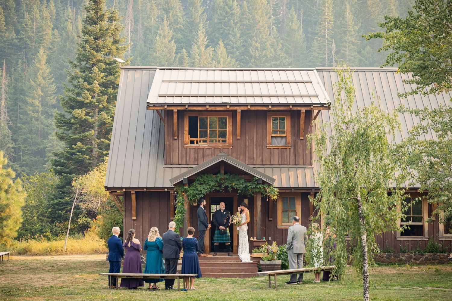 Wedding guests gather outside a rustic wooden lodge with metal roof in a mountain forest setting.