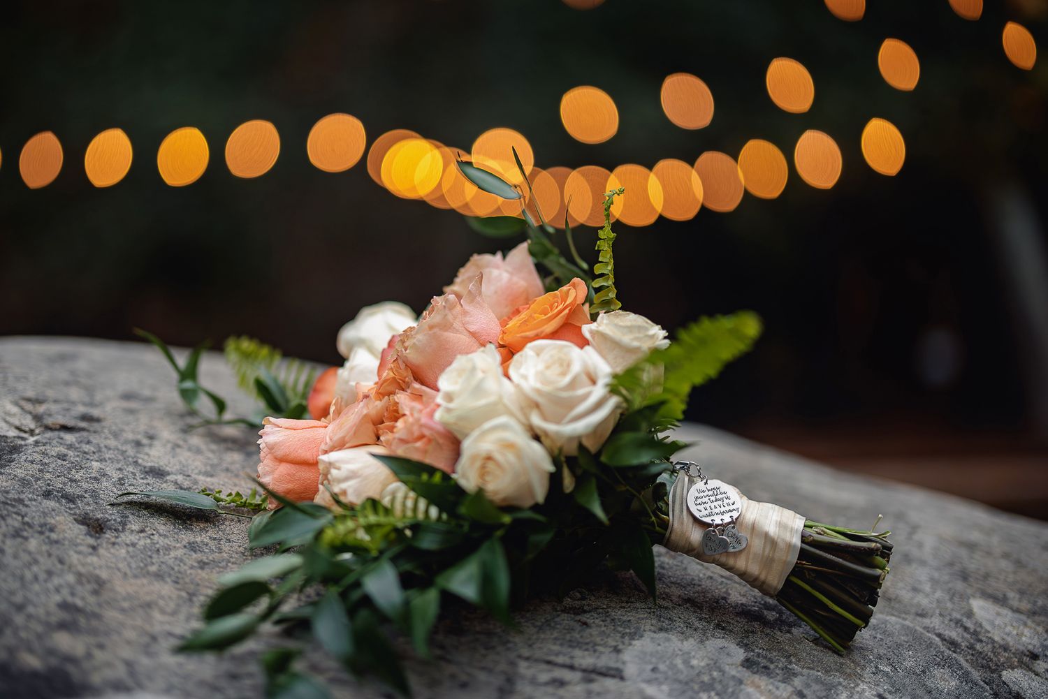 A wedding bouquet of coral and white roses with greenery rests on rustic wood surface under glowing string lights.
