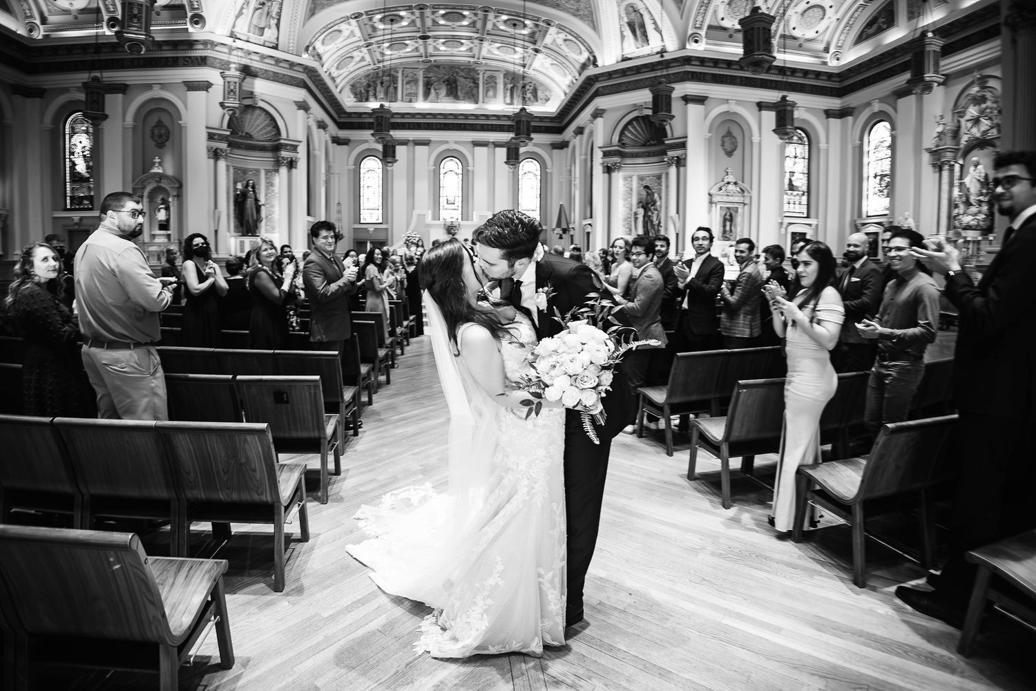 A couple walks down the aisle of an ornate church with high ceilings and traditional wooden pews filled with wedding guests.