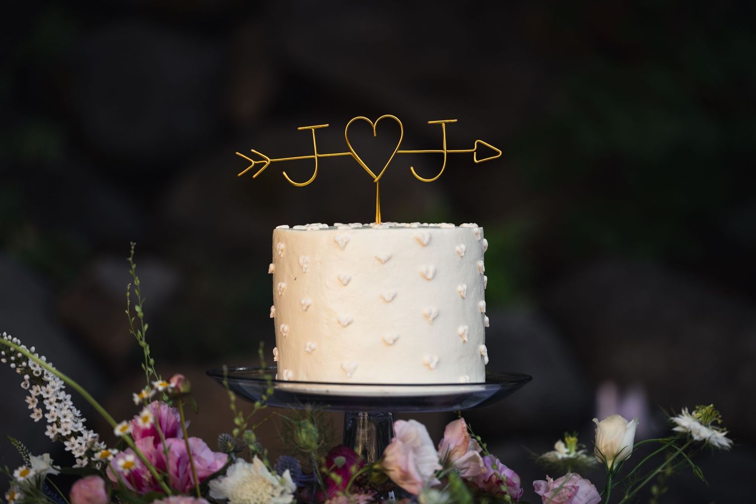 A white wedding cake with dotted texture and gold arrow heart topper sits above pink and purple flowers on a dark stand.