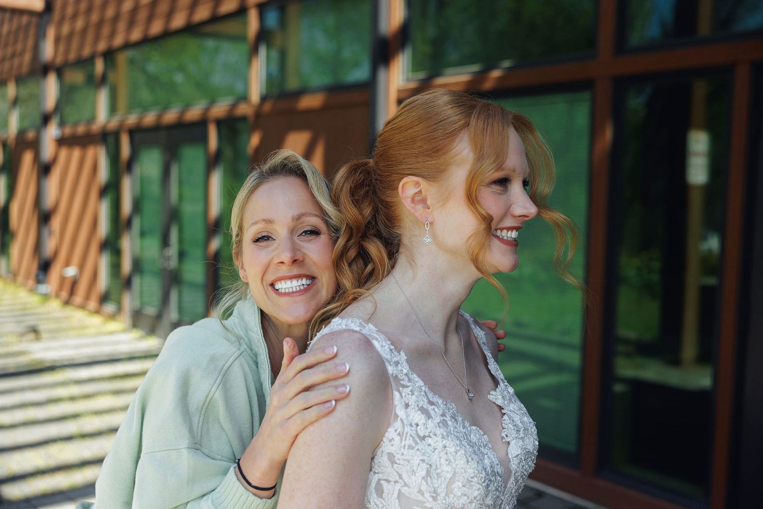Bride in lace gown smiling with bridesmaid in mint jacket outdoors near a rustic building on wedding day.