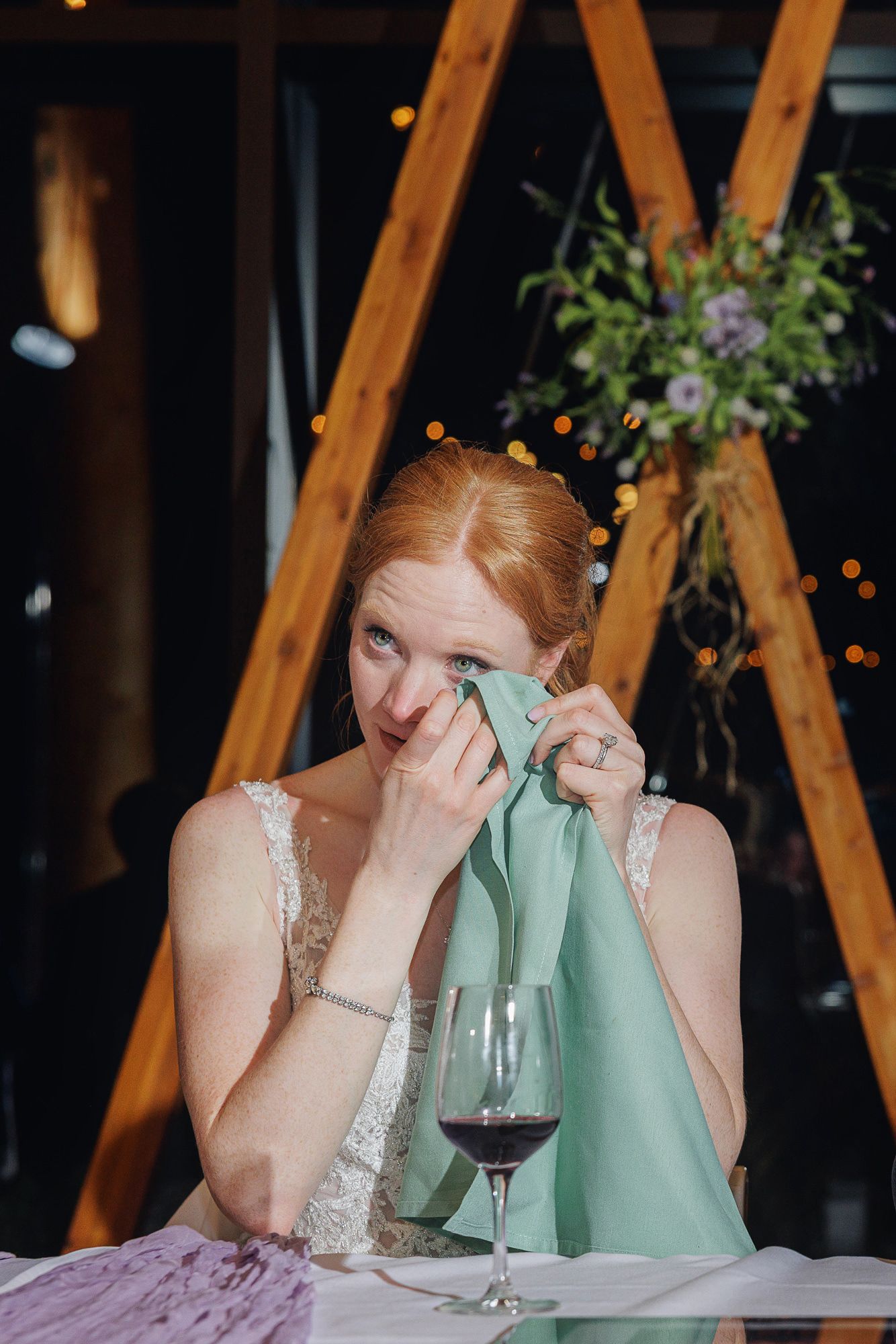Emotional bride in white lace dress wiping tears with green napkin at wedding reception table with red wine glass.