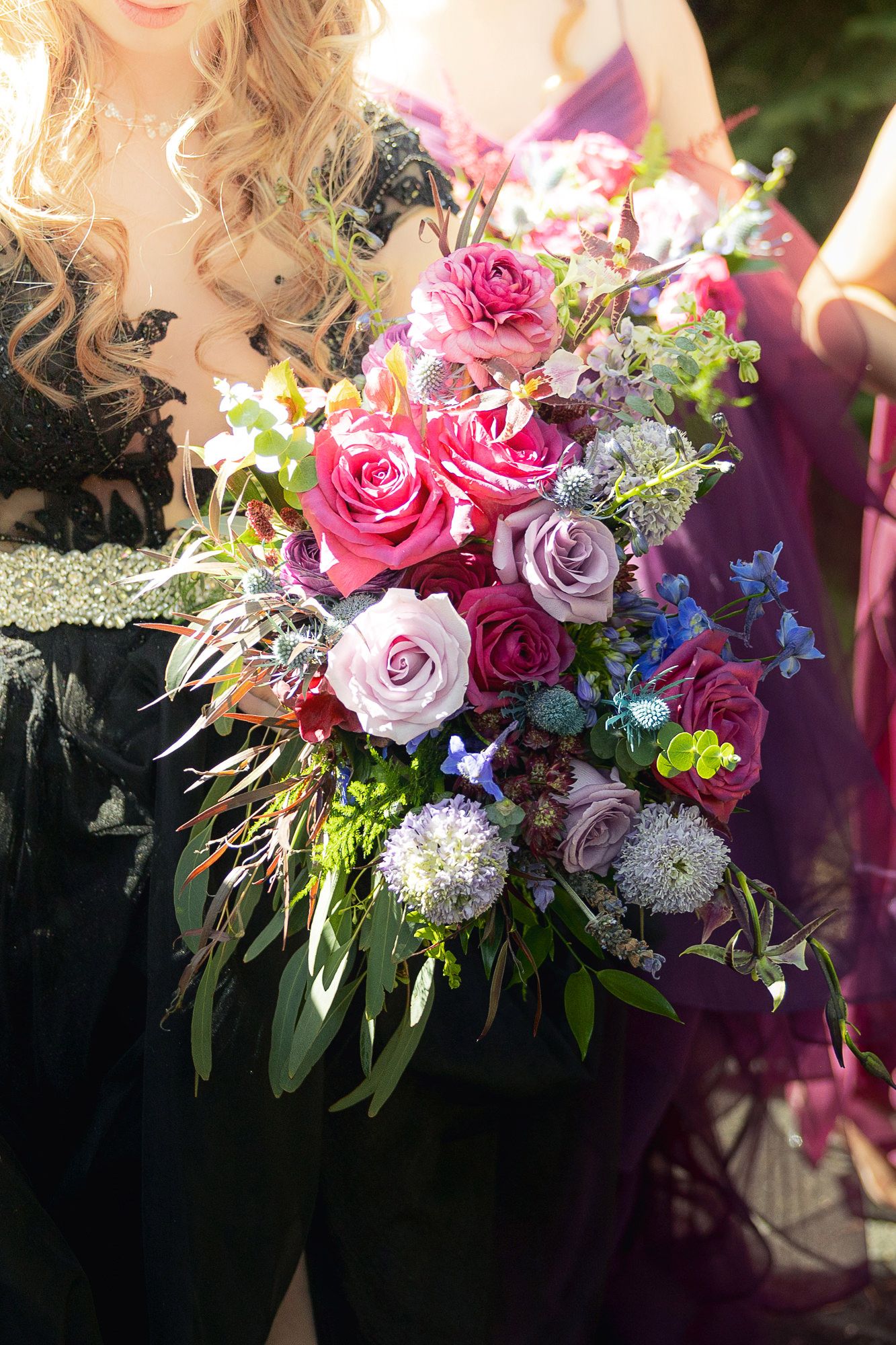 Woman in black dress holding a lush bridal bouquet of pink, purple roses, blue flowers, and greenery in sunlight.