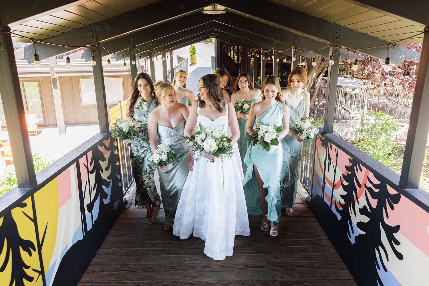 Bride and bridesmaids in mint green dresses walk through a colorful covered bridge holding white floral bouquets.