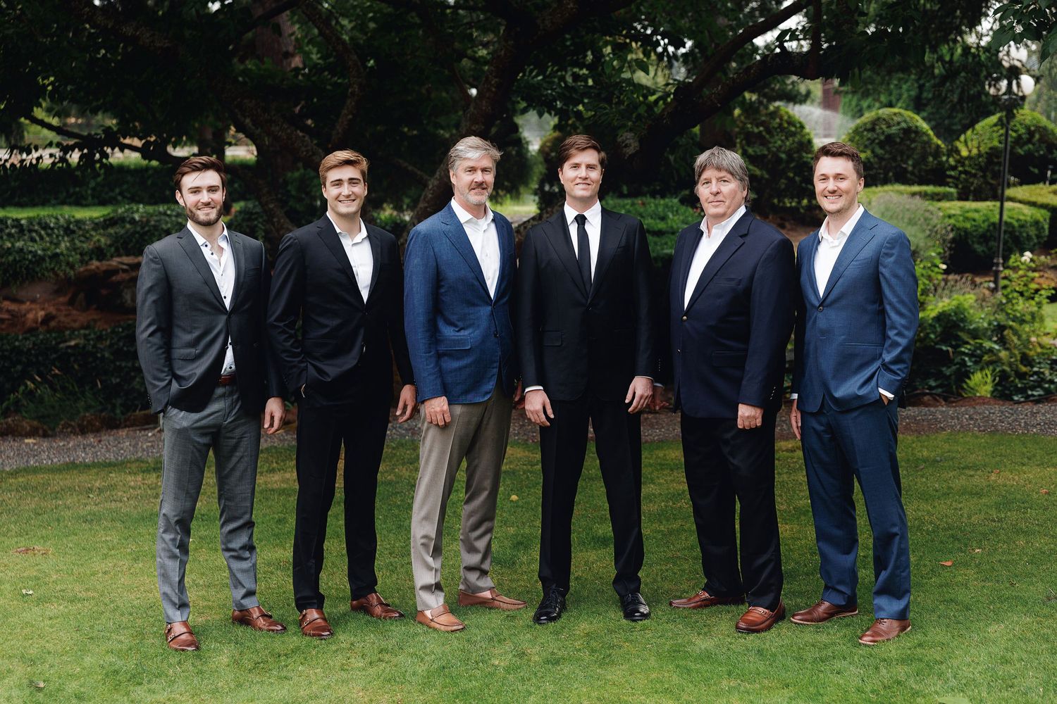 Six well-dressed men in suits posing together outdoors on a green lawn with lush garden hedges in the background.