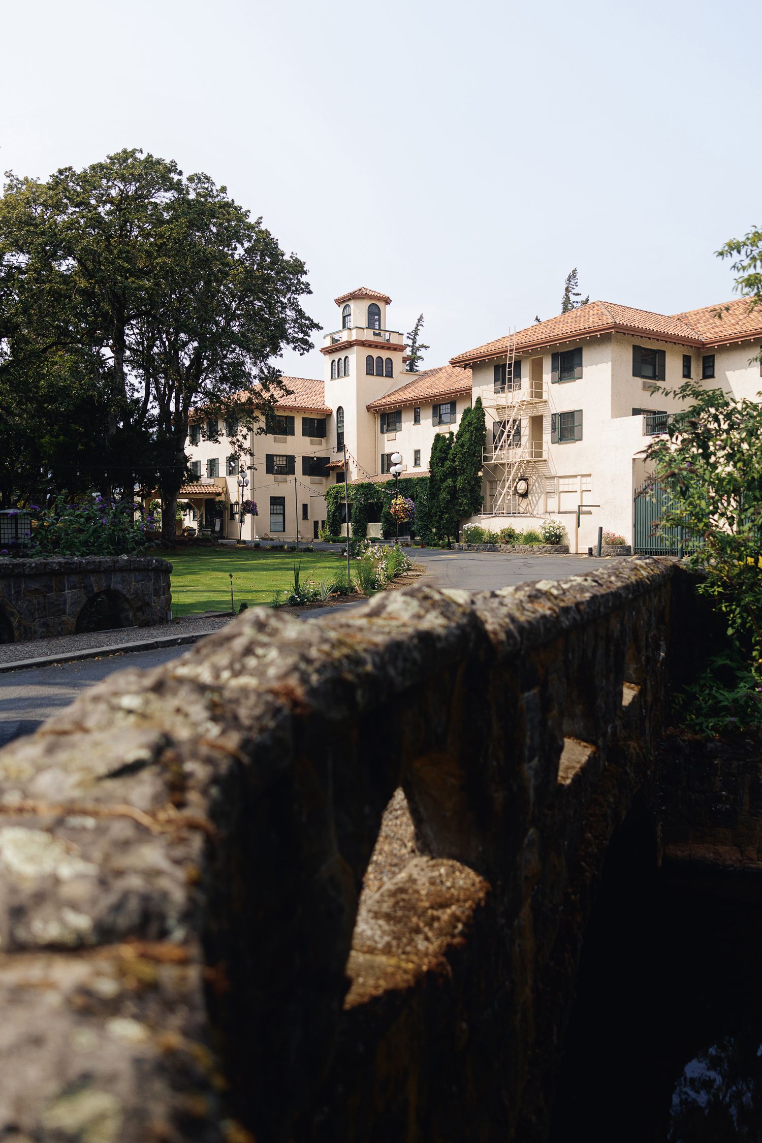 Historic Italian-style villa with stone bridge arch, terracotta roof, and clock tower surrounded by lush trees.