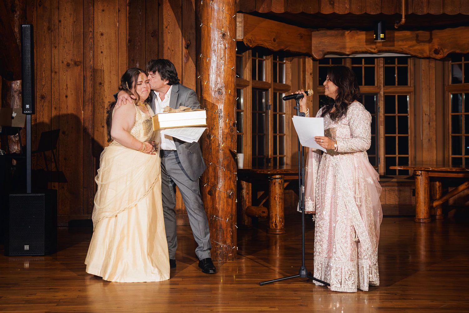 Couple sharing a kiss at rustic log cabin wedding reception while officiant reads from notes at microphone.