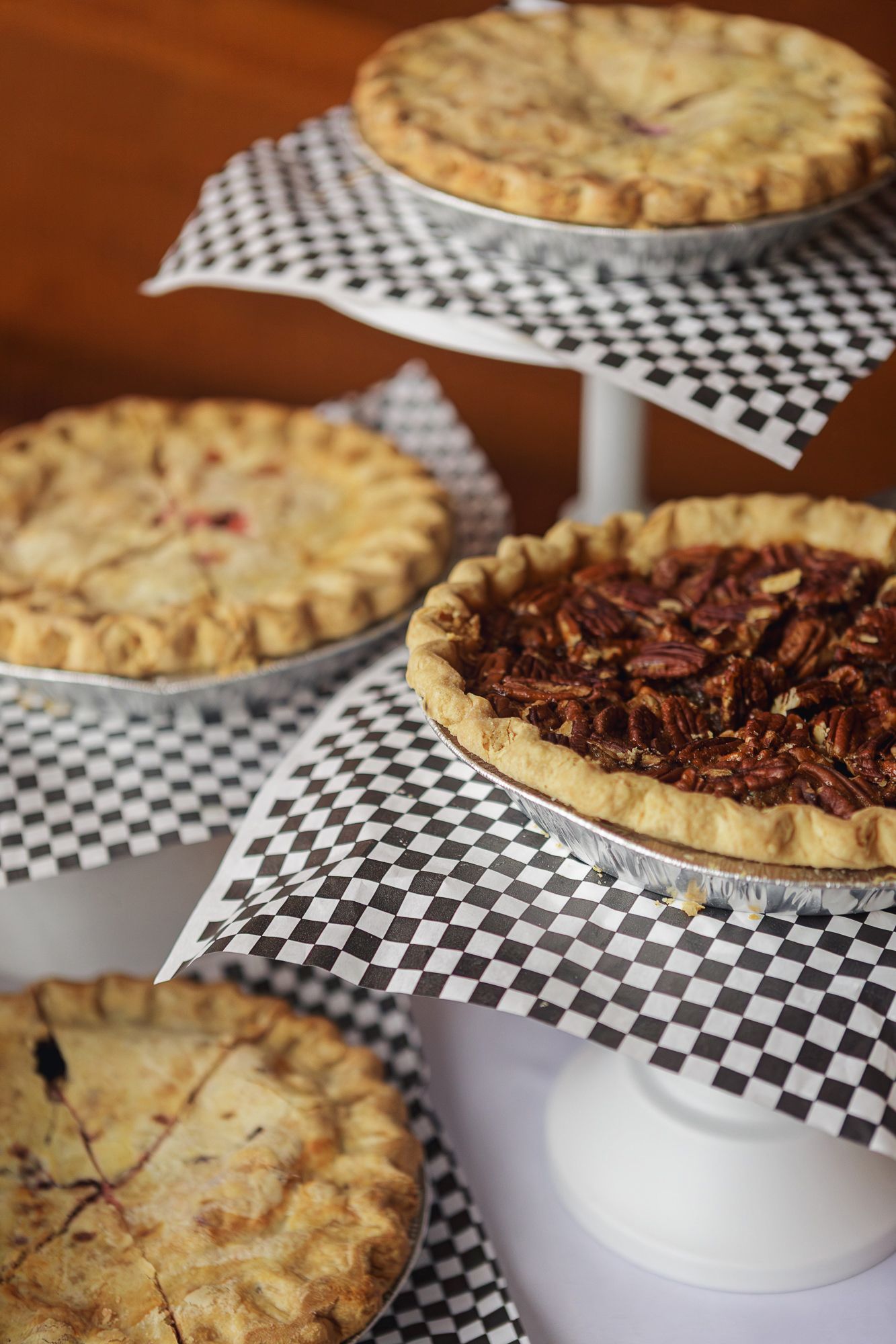 An assortment of homemade pies with flaky crusts displayed on white cake stands with checkered paper liners.