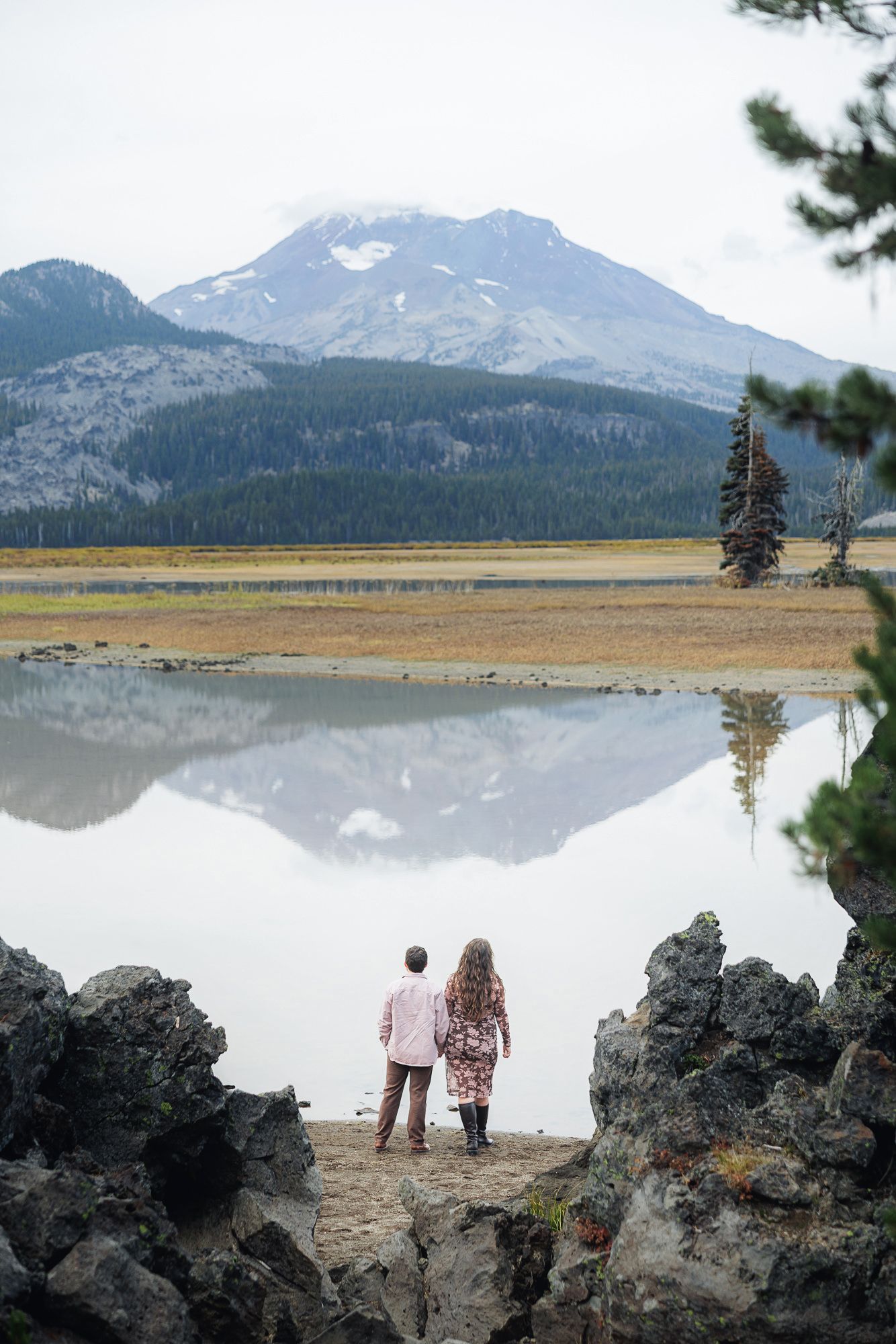 Scenic panoramic view of a mountain lake with volcanic rocks in the foreground and snow-capped peaks reflecting in calm waters.