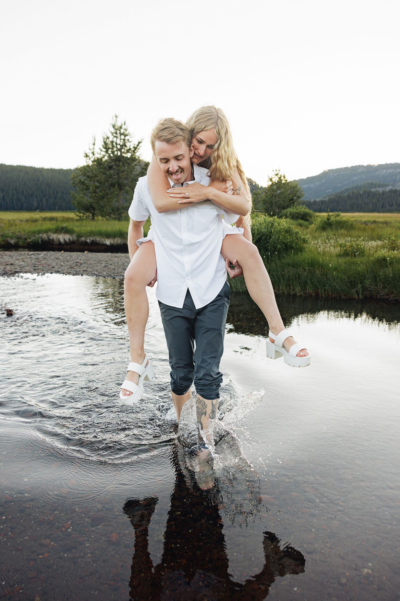 A young man carries a smiling blonde woman piggyback through a shallow stream in a scenic meadow.