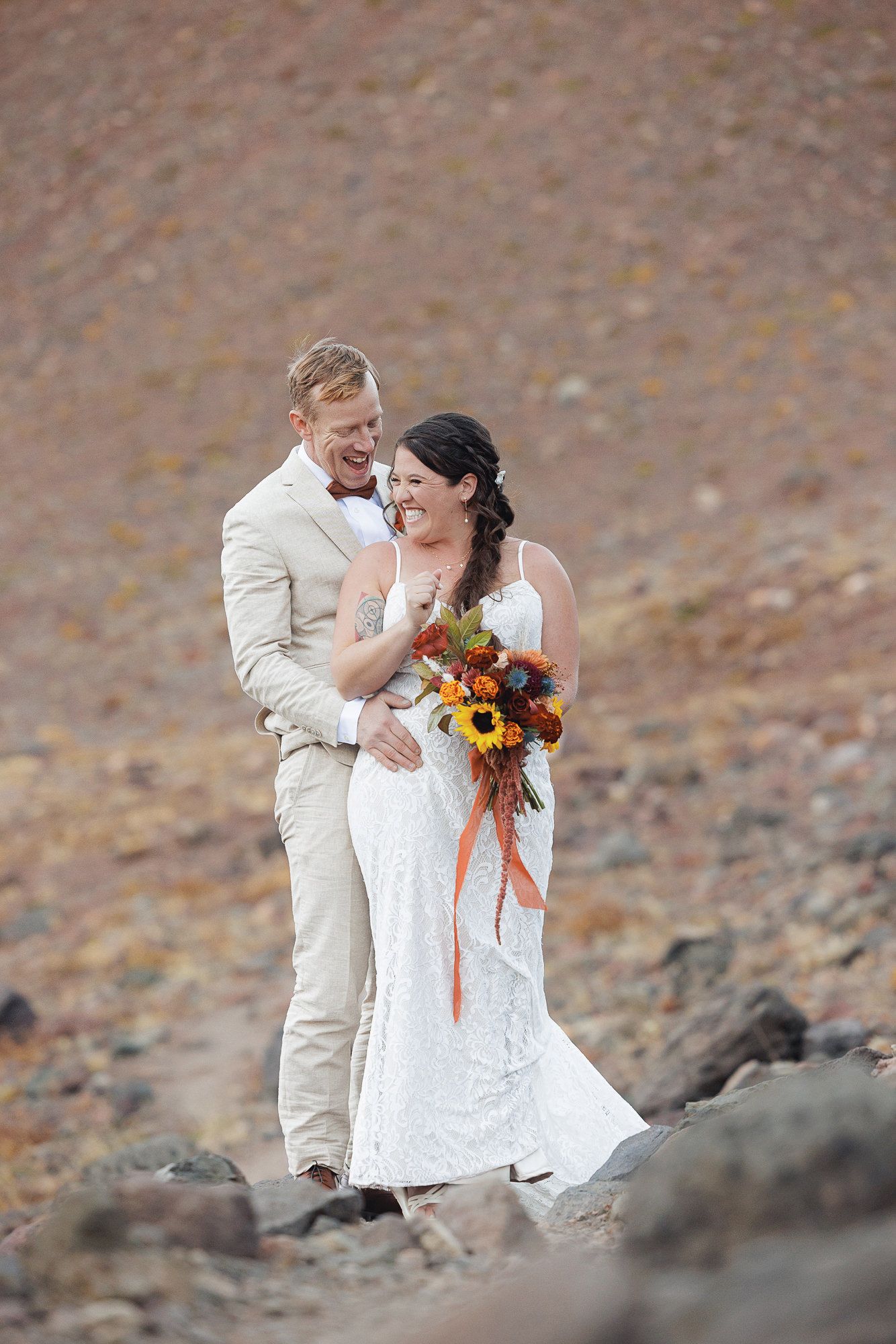 A smiling bride in a white lace gown holds a sunflower bouquet while her groom in a tan suit embraces her on a rocky mountain landscape.
