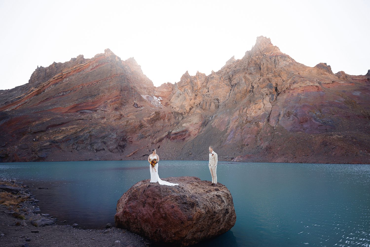 Two people in white stand on a large boulder in a calm turquoise mountain lake, with rugged red rocky peaks rising behind them.
