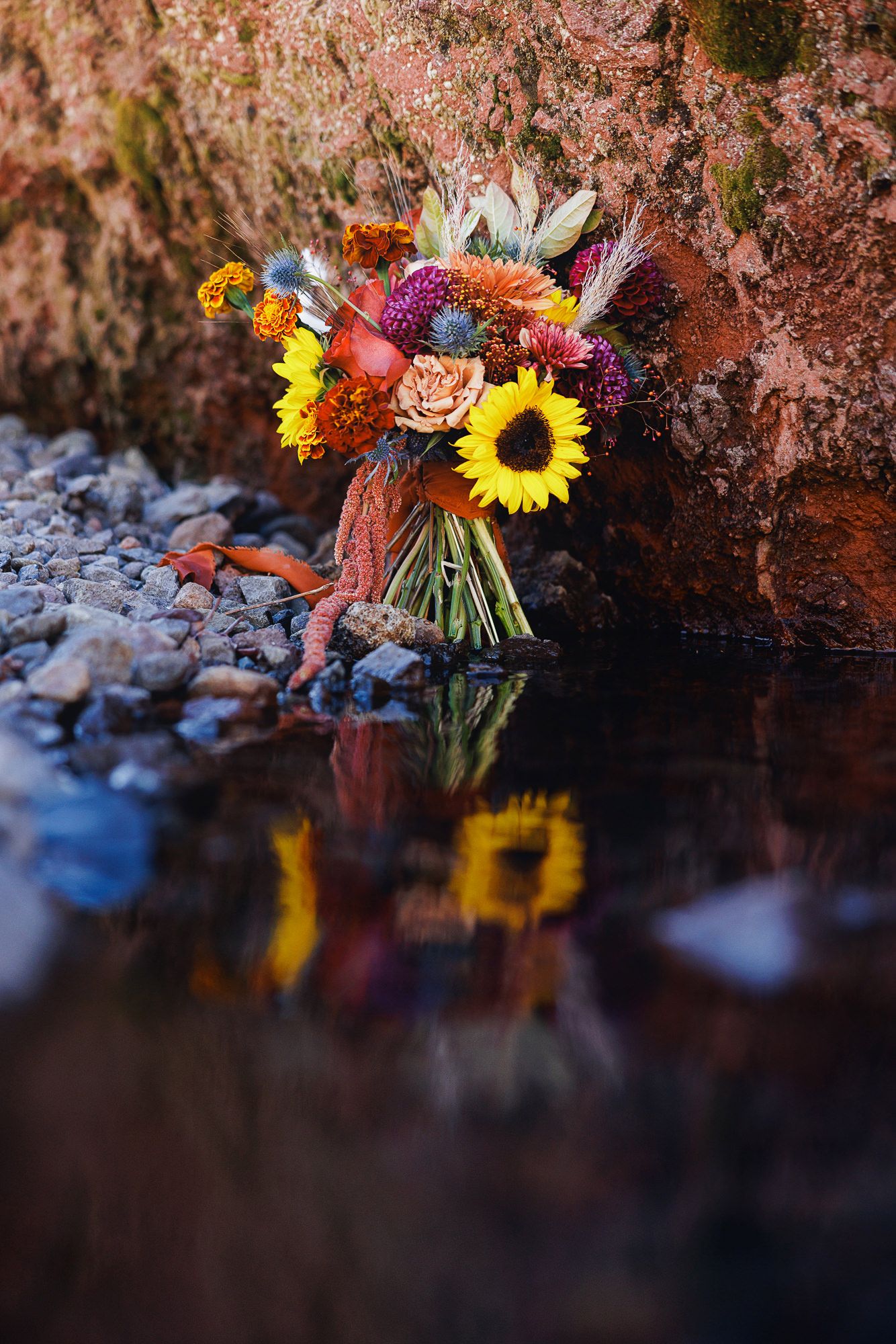 Colorful autumn wedding bouquet with sunflowers, dahlias and thistles resting on rocks beside still water, reflected in the surface.