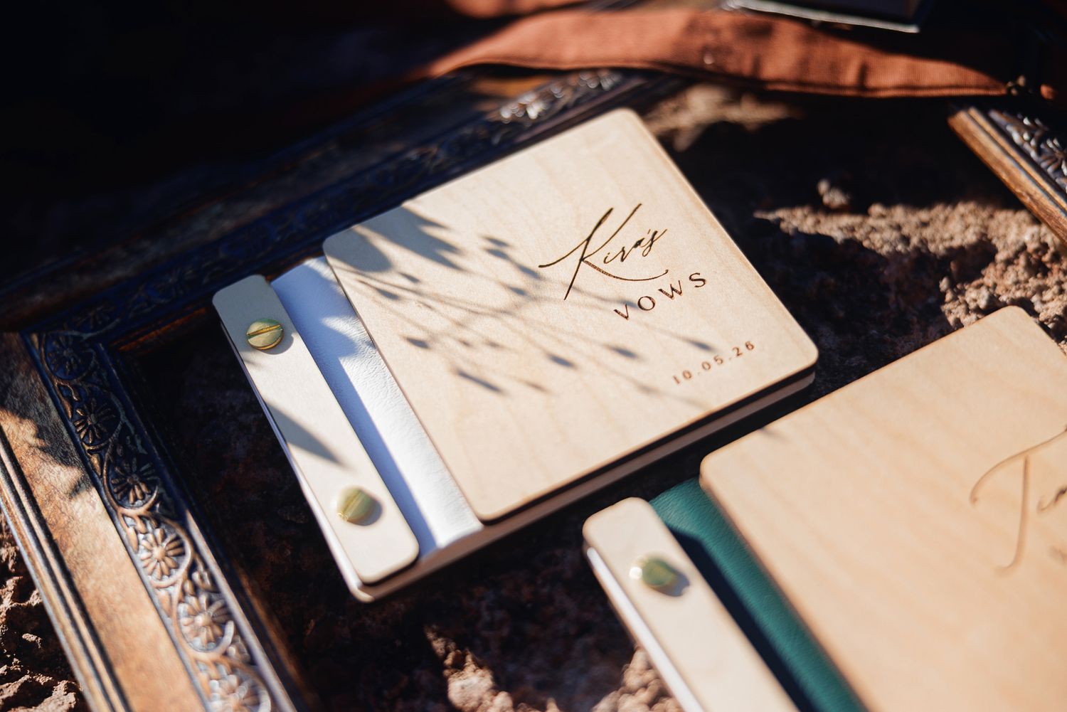 Personalized wooden wedding vow books with engraved script titles and gold snap closures resting in an ornate vintage frame on sandy ground.