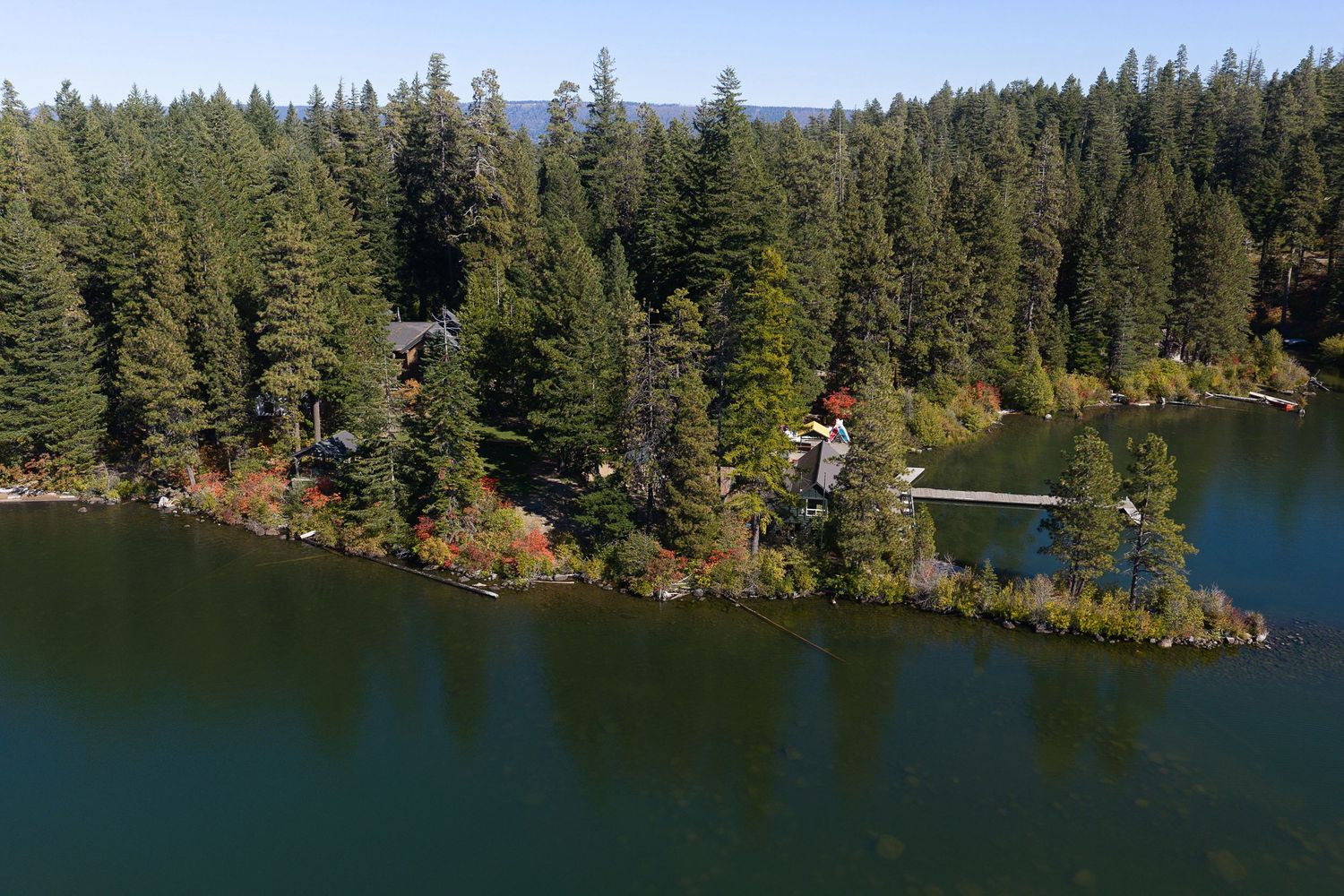 Aerial view of a lakeside property surrounded by dense evergreen forest with a private dock extending into calm blue water.