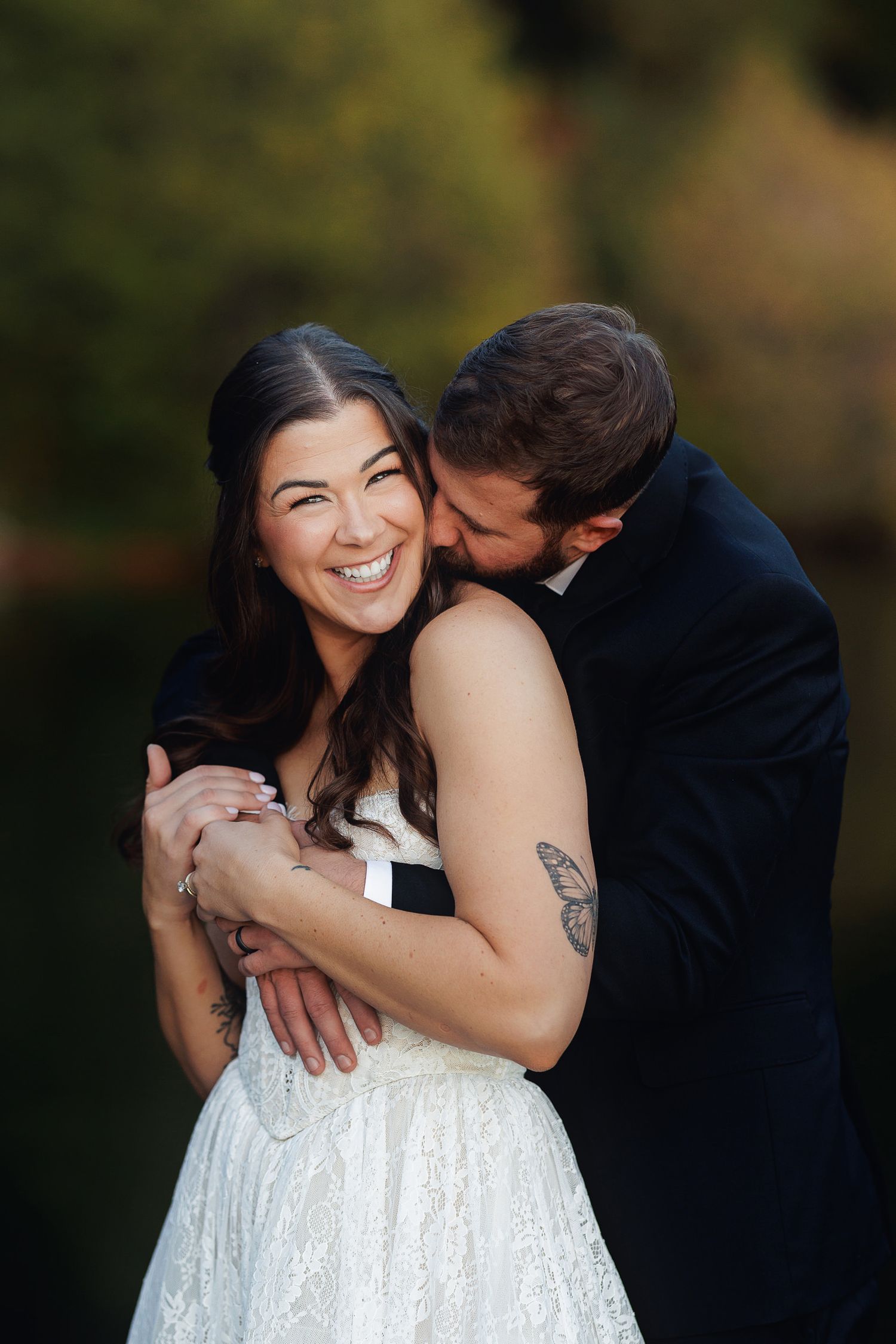 Bride in lace dress laughs joyfully as groom in navy suit embraces and kisses her cheek outdoors.