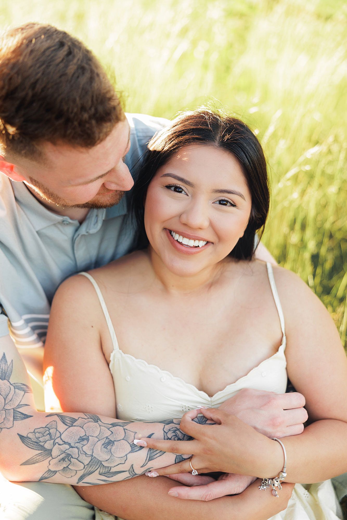 Couple embracing in a sunlit field, woman smiling brightly, man with floral tattoo hugging her from behind.
