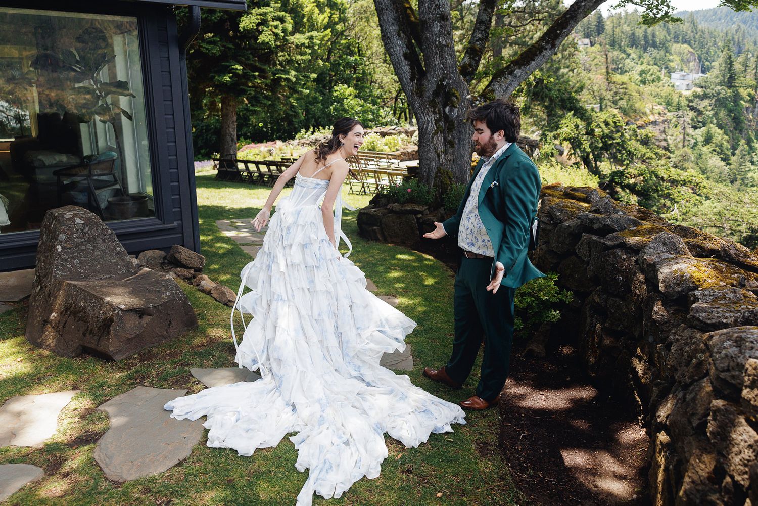 Bride in flowing white gown and groom in teal suit share a first look moment outdoors near stone wall and lush trees.