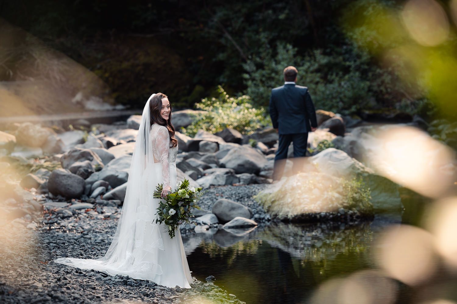 A bride in a white dress and groom standing on a rocky riverbank among trees in an artistic wedding photo.