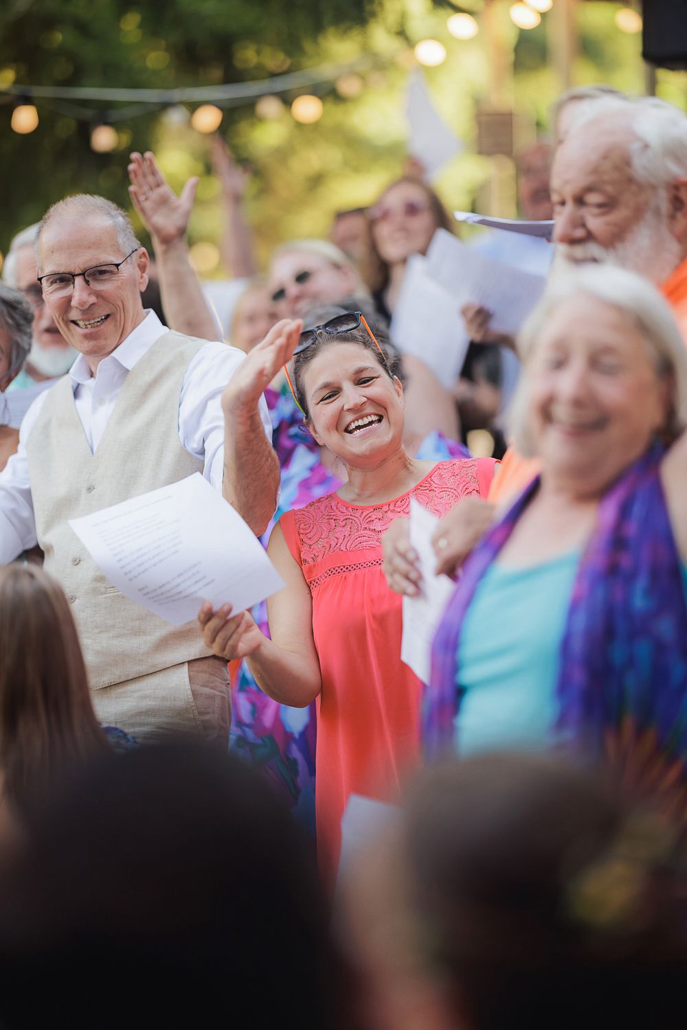 A cheerful crowd of people enjoying a festive outdoor celebration in warm evening light with string lights overhead.