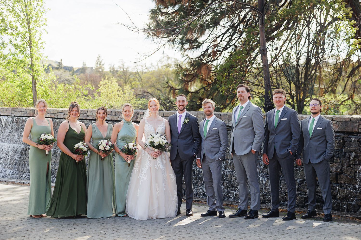 Wedding party posing outdoors with bridesmaids in sage green gowns and groomsmen in gray suits beside a waterfall.