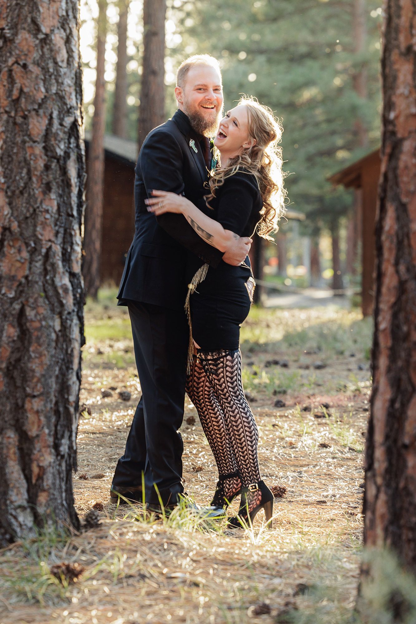 Couple embracing and laughing together among tall pine trees, both dressed in black outfits in a forest setting.