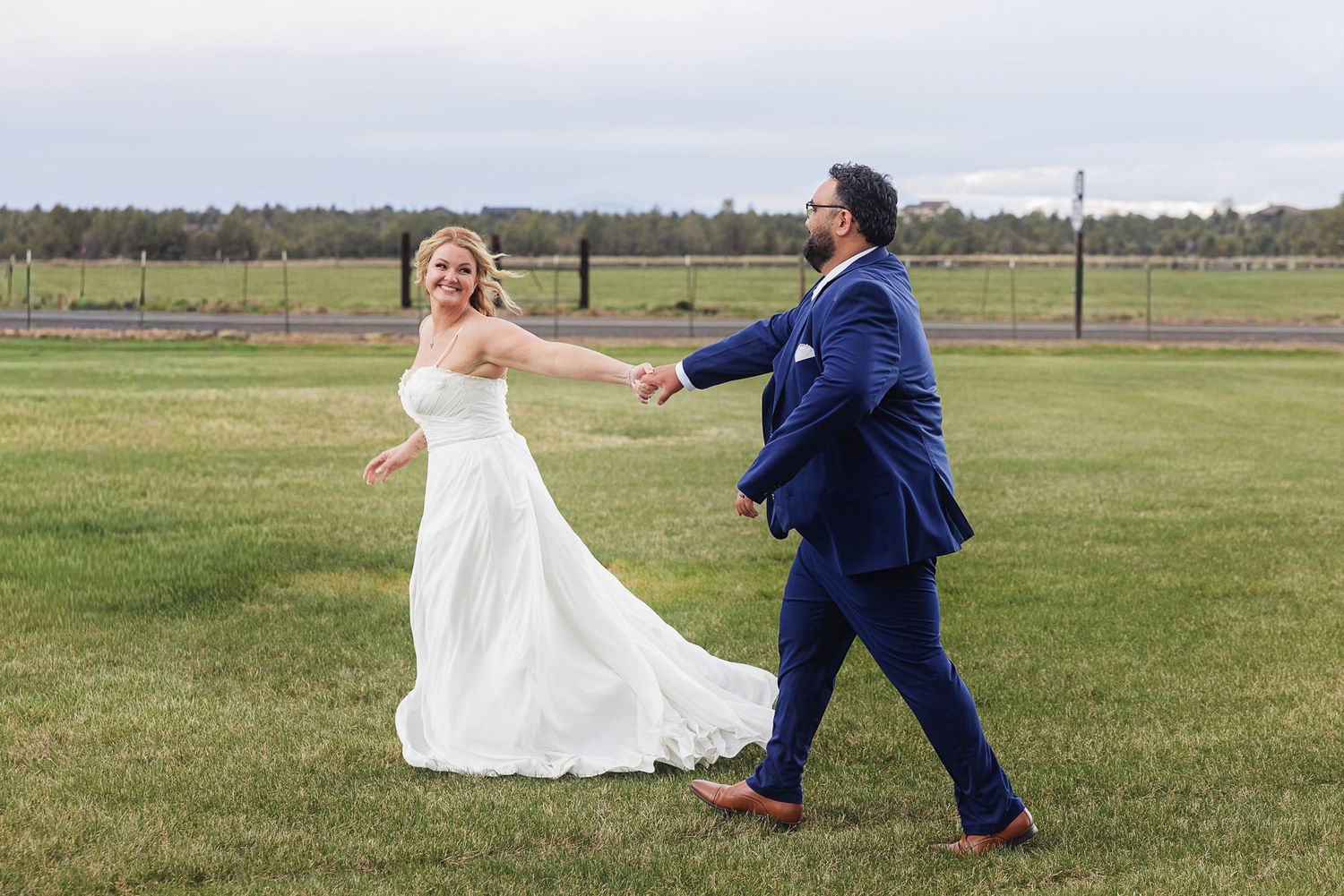 Bride in white gown and groom in blue suit walk hand in hand across green lawn at outdoor wedding venue.