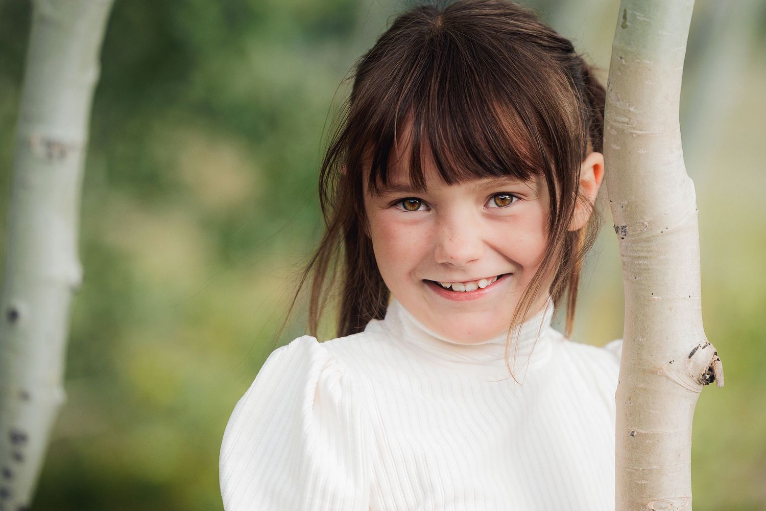 Portrait of young person in white sweater smiling brightly against birch tree.