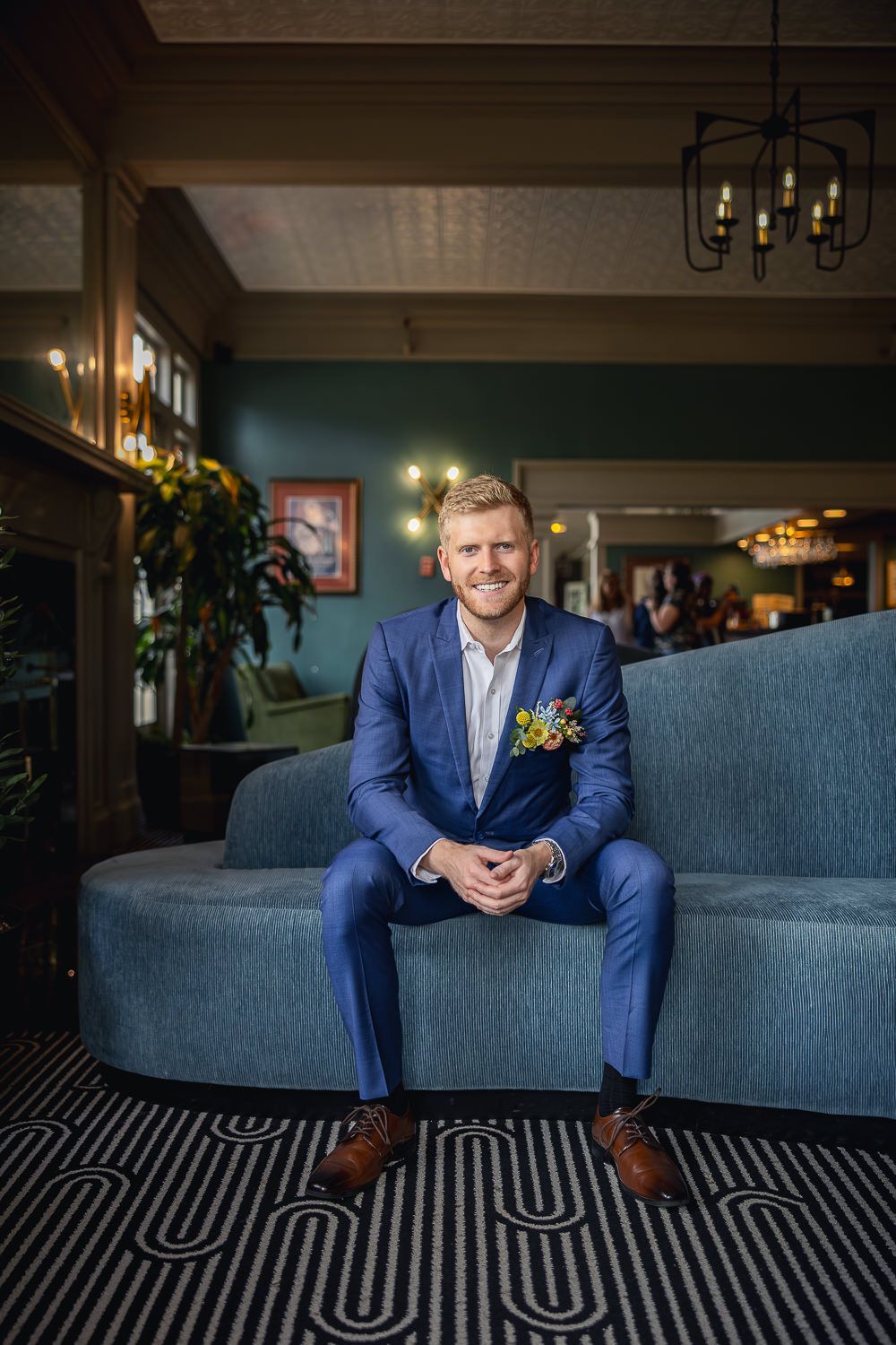 A groom in a fitted navy suit sits on a blue velvet couch in an elegantly decorated room with dark walls and chandeliers.