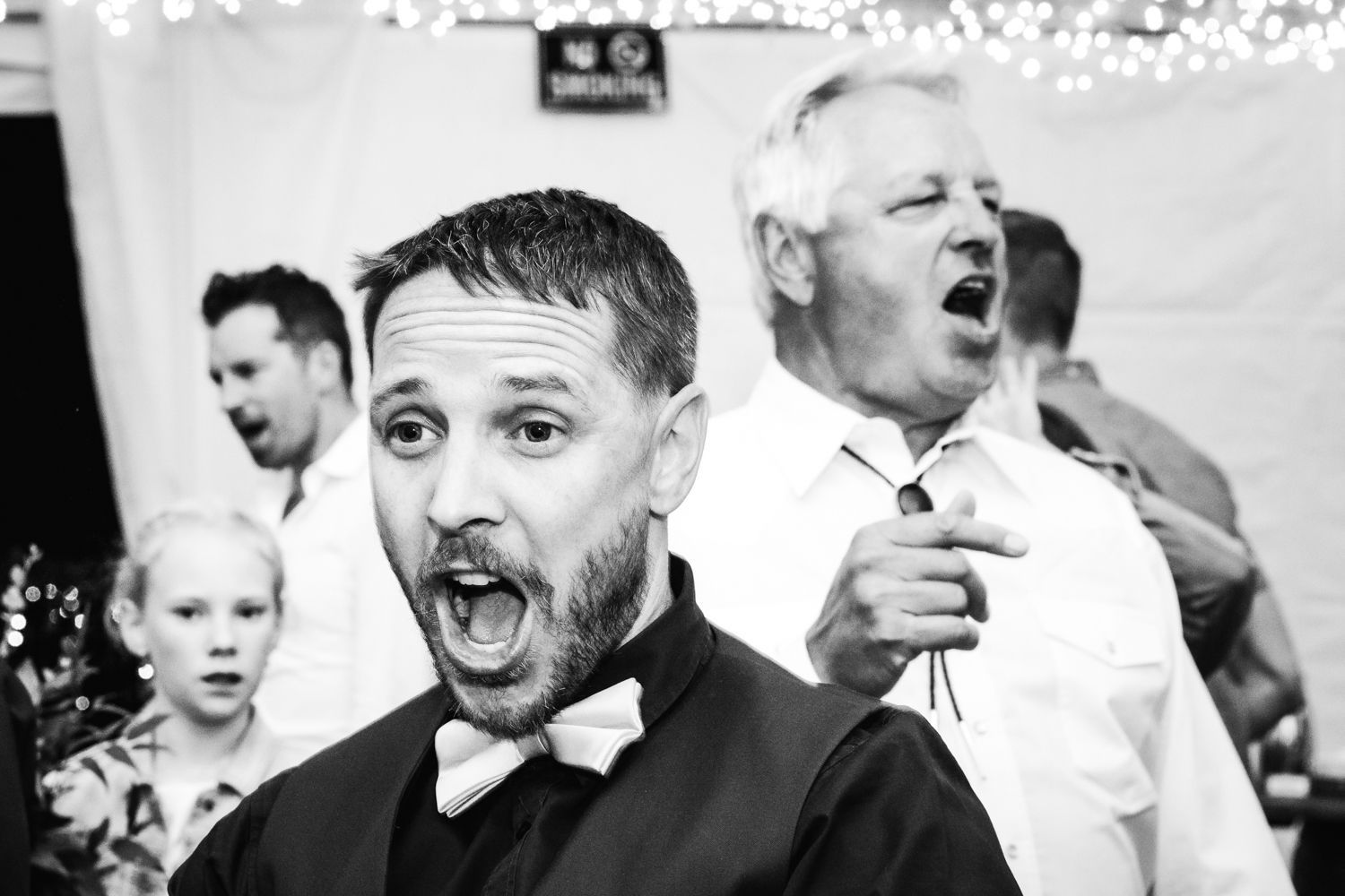 A black and white candid image showing the excited and animated expressions of wedding guests during a celebration moment.