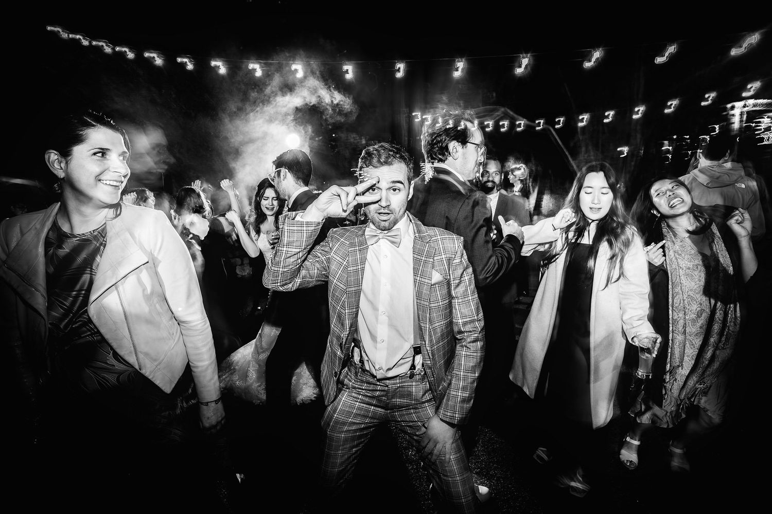 Black and white photo showing people dancing and celebrating at a lively party under string lights.