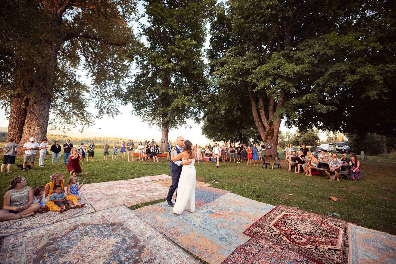 Wedding ceremony takes place on vintage rugs laid out on grass with guests sitting around under shady trees.